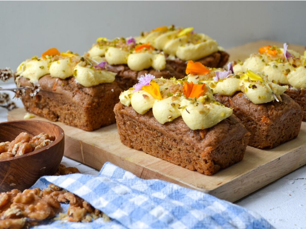 Three rectangular vegan carrot cake loaves with yellow icing, pistachios, and edible flowers on a board near walnuts and a blue cloth.