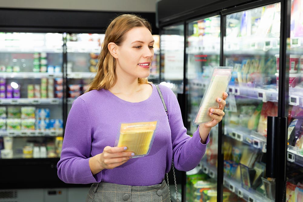 A woman holds two different varieties of cheese in front of the refrigerated section of the supermarket. 