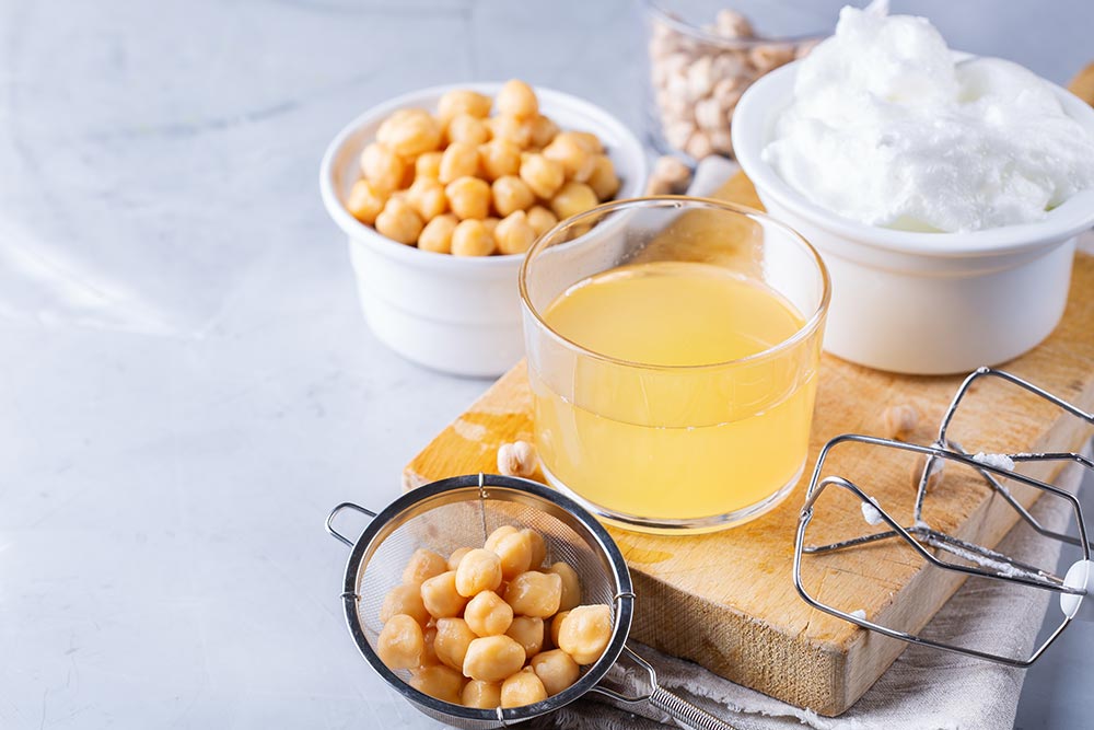 A bowl of drained chickpeas sit beside a wooden board with a small glass of aquafaba, and a small bowl of aquafaba whip.