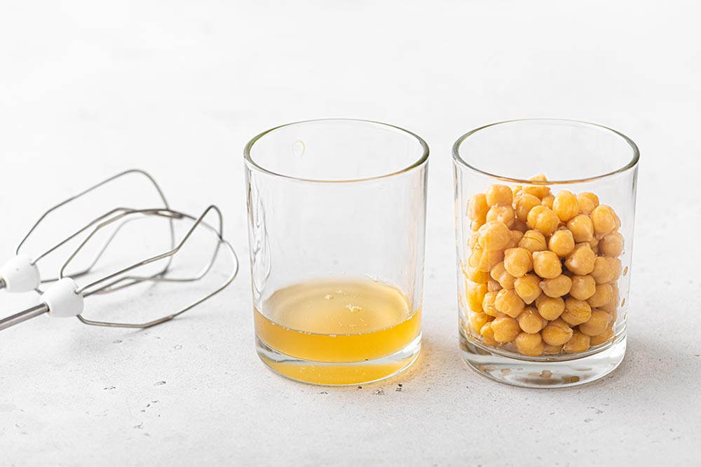 A pair of beaters sit on a white bench with a glass with a small amount of aquafaba next to them, and another glass full of drained chickpeas.