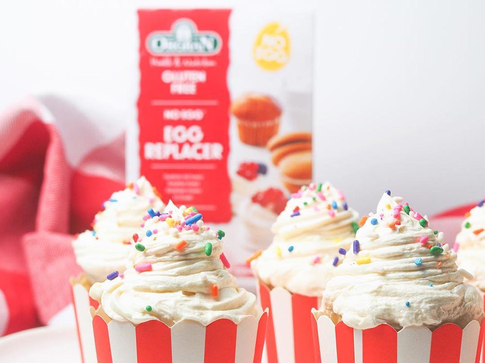 Cupcakes in red and white striped patty pans are topped with a white frosting and coloured sprinkles. In the background, slightly blurred, there is a packet or Orgran No-Egg Egg Replacer. 