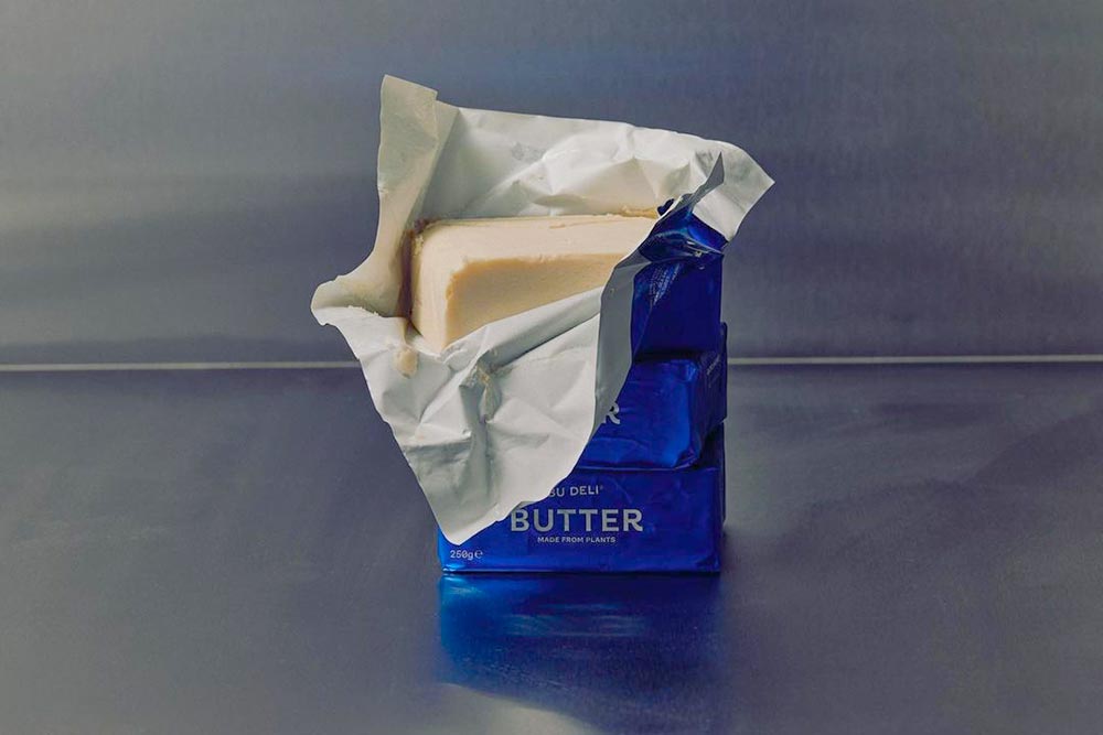 A block of butter stands up on a metal bench with its blue packaging partially opened, revealing a butter slab.