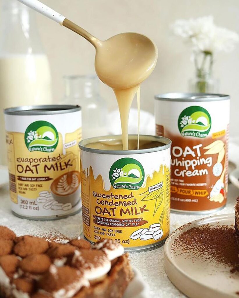 Three tins of different oat baking products sit on a bench with the middle tin of sweetened condensed oat milk open. A spoon is being held above it, dripping a stream of sweet, sticky liquid back into the tin.