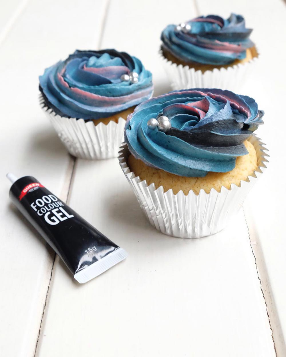 Three cupcakes in silver patty pans sit atop a white surface, with a dark blue to purple swirled frosting decorated with tiny silver cachous. A tube of food colour gel sits open beside them. 