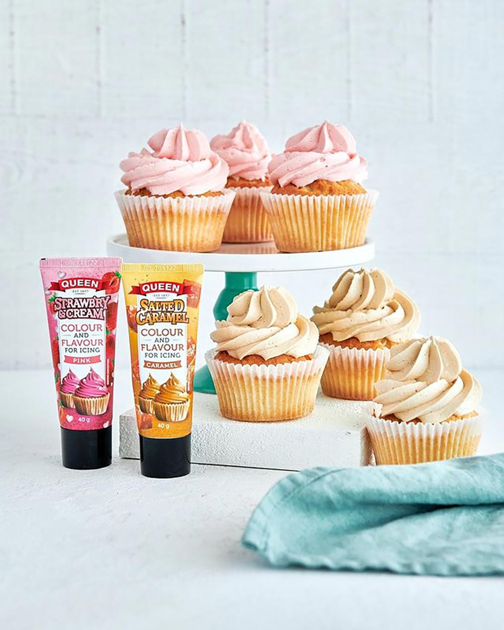 A cupcake stand holds pink frosted cupcakes, while caramel frosted cupcakes sit below. Two tubes of colour and flavouring icing gel stand beside them.