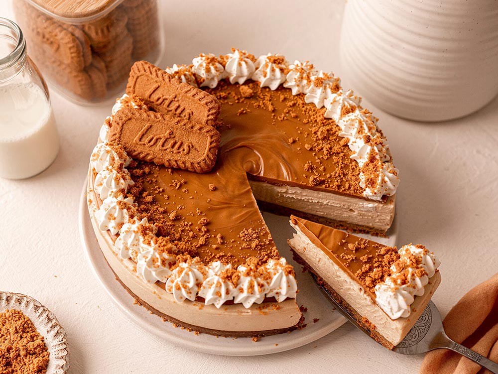 A hand holding a cake server pulls a slice of Biscoff cheesecake away from the full cake. The cake looks beautifully creamy and is decorated with full Biscoff cookies, as well as cookie crumbles.