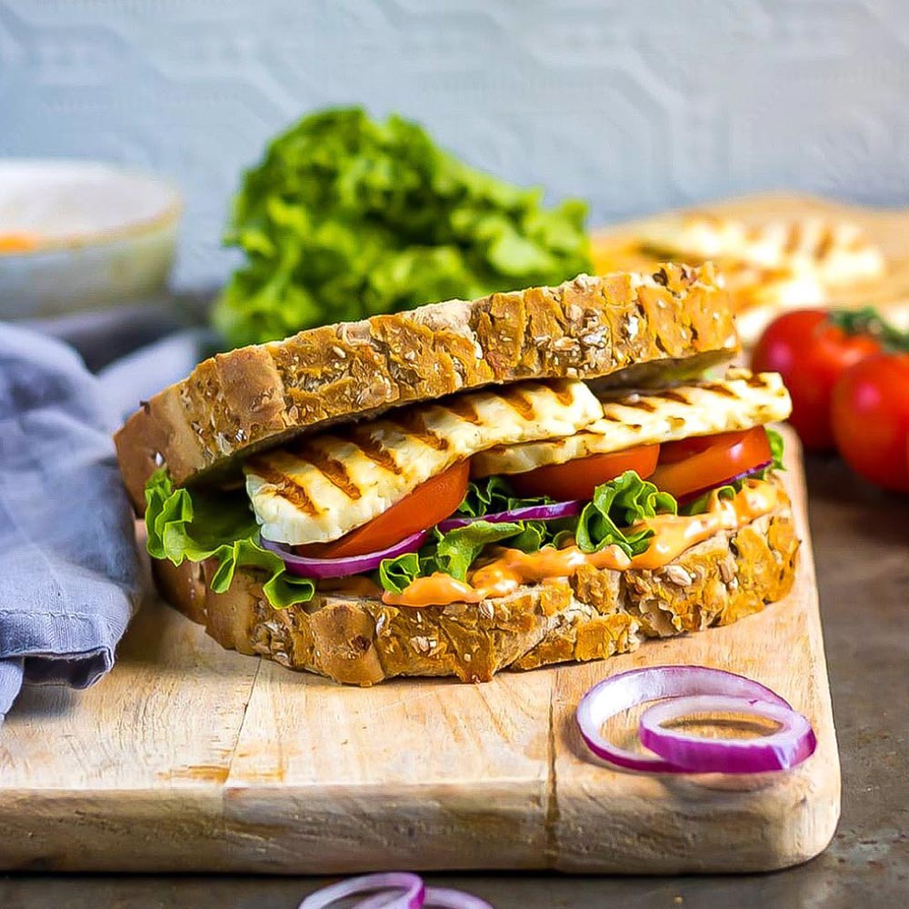 A sourdough toastie sits on a wooden chopping board. It is slightly open to reveal two generous slices of a grilled halloumi.