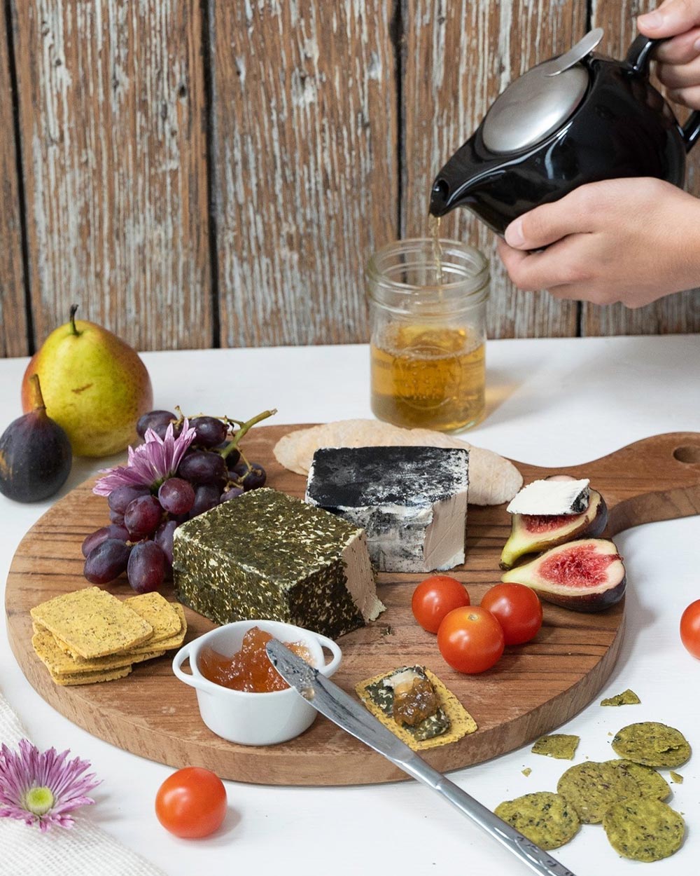 A charcuterie board features two different style of chevre form The Vegan Dairy. It's beautifully decorated with grapes, an assortment of crackers, figs, tomatoes, and a quince paste.