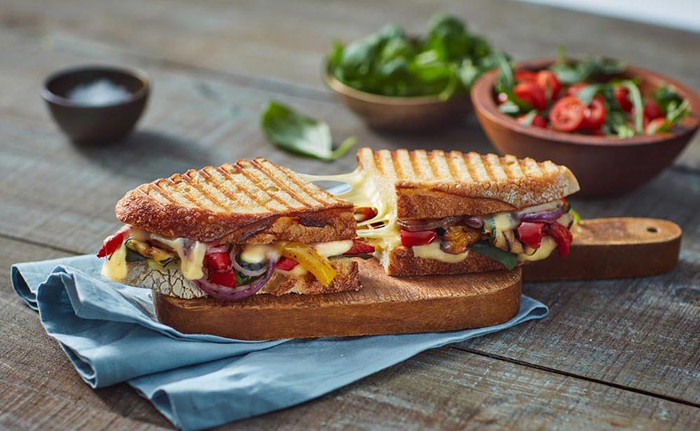 A grilled sourdough toastie packed with roast veg is served on a wooden serving board. It has been cut in half and separated slightly to show how stretchy, gooey, and melty the plant-based cheese is. 