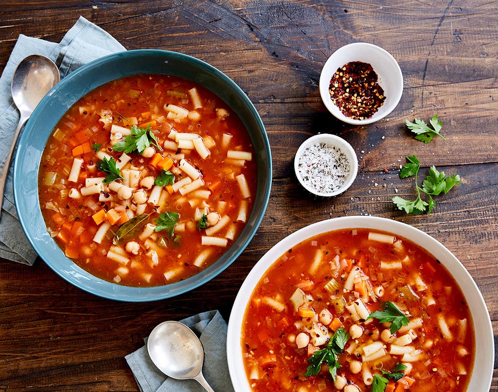 Overhead view of two bowls of rich tomato soup, laden with macaroni and chickpeas. They're both garnished with parsley.