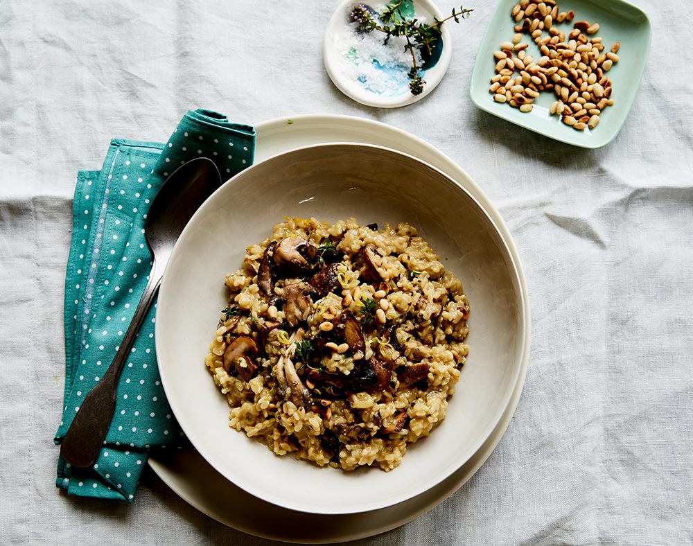 A bowl of hearty-looking mushroom risotto sits on a table ready to eat. It's topped with crunchy pine nuts, with more in a small bowl to the side.