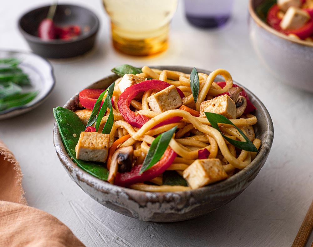 A bowl of hokkien noodles served with stir-fried snow peas, peppers, greens, and crispy cubes of tofu.