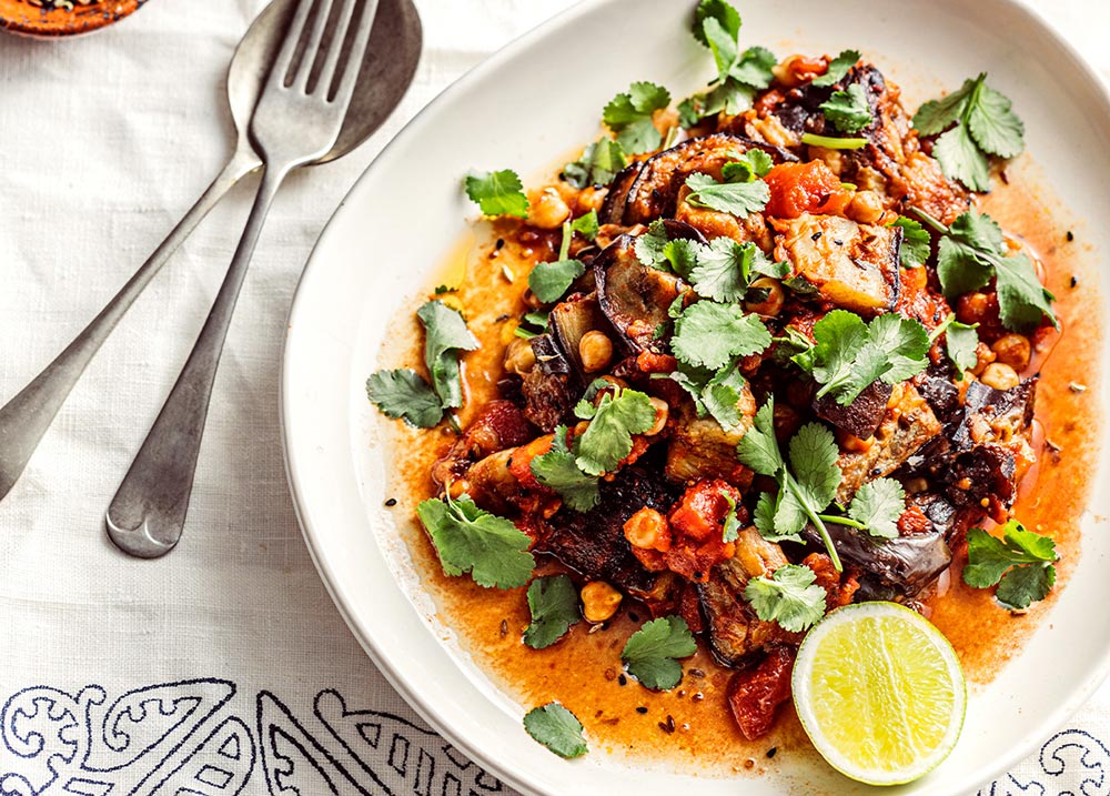 A plate is piled high with eggplant and chickpea curry, garnished with herbs and served with a lemon slice.