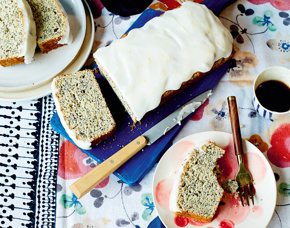 A loaf of lemon and poppy seed cake with a sweet white icing. A few slices have been cut and sit on nearby plates. One slice has a bite taken out. 