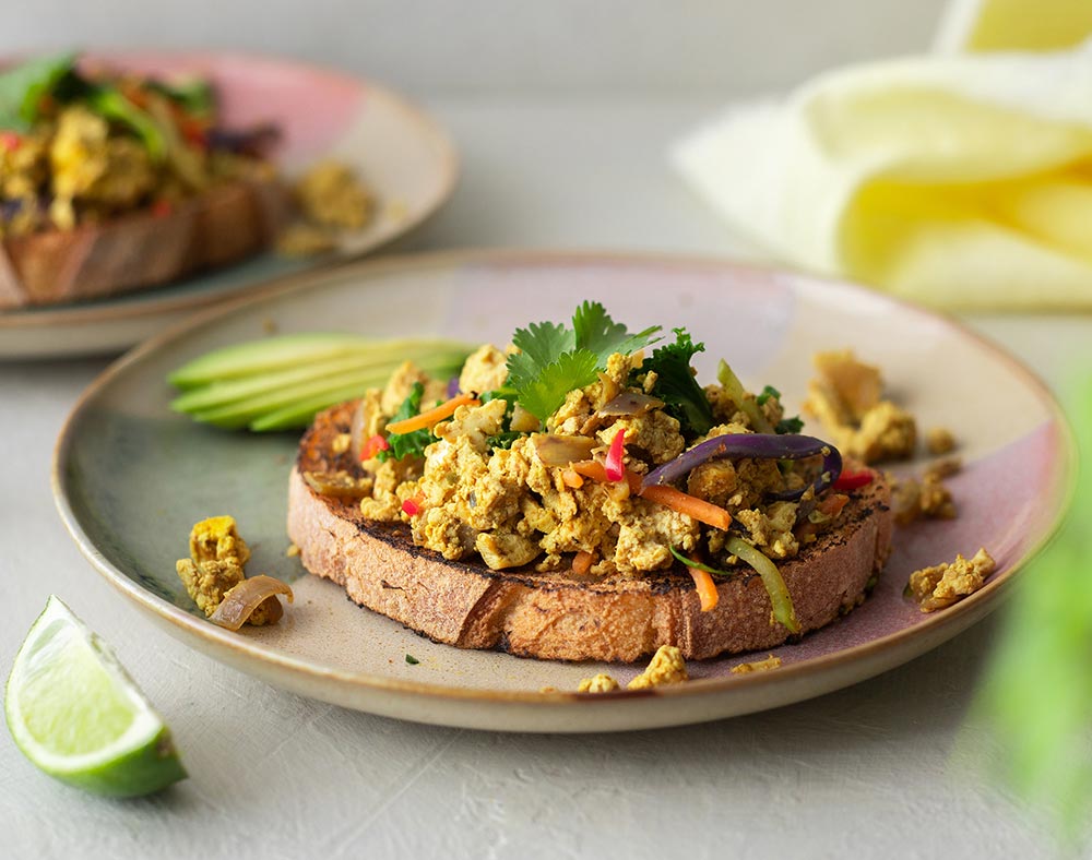 A slice of toast topped with smoky tofu scramble. It's garnished with fresh greens and a wedge of lime sits to the side.