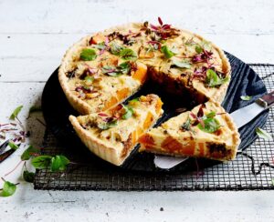 An eggy looking pumpkin quiche-style tart sits on a wire rack to cool. Two slices have been cut to show the inside. The slices are packed with veg.