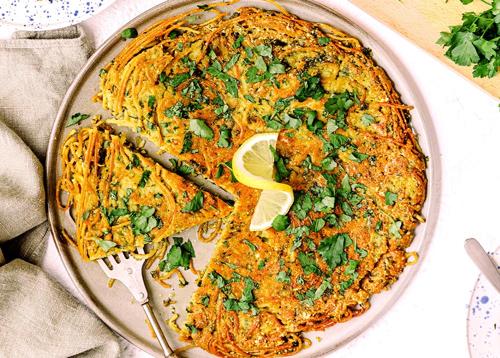 An overhead view of a spaghetti frittata garnished with fresh greens and a slice of lemon. 