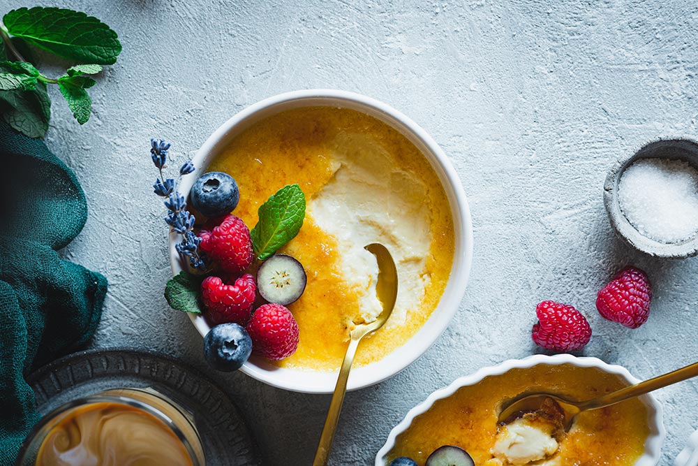 Overhead view of a creme brulee topped with fresh berries. A spoon has already cracked the shell and has started on the sweet custard below. 