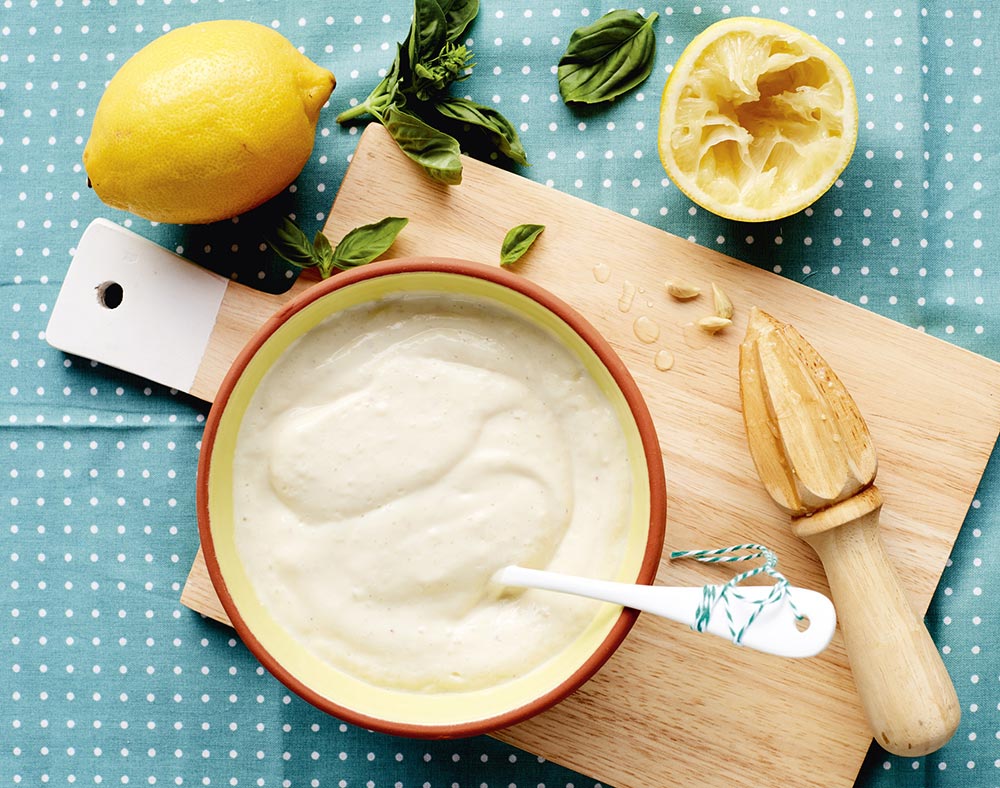 A bowl of mayo sits on a wooden chopping board, with lemon and basil scattered around it.