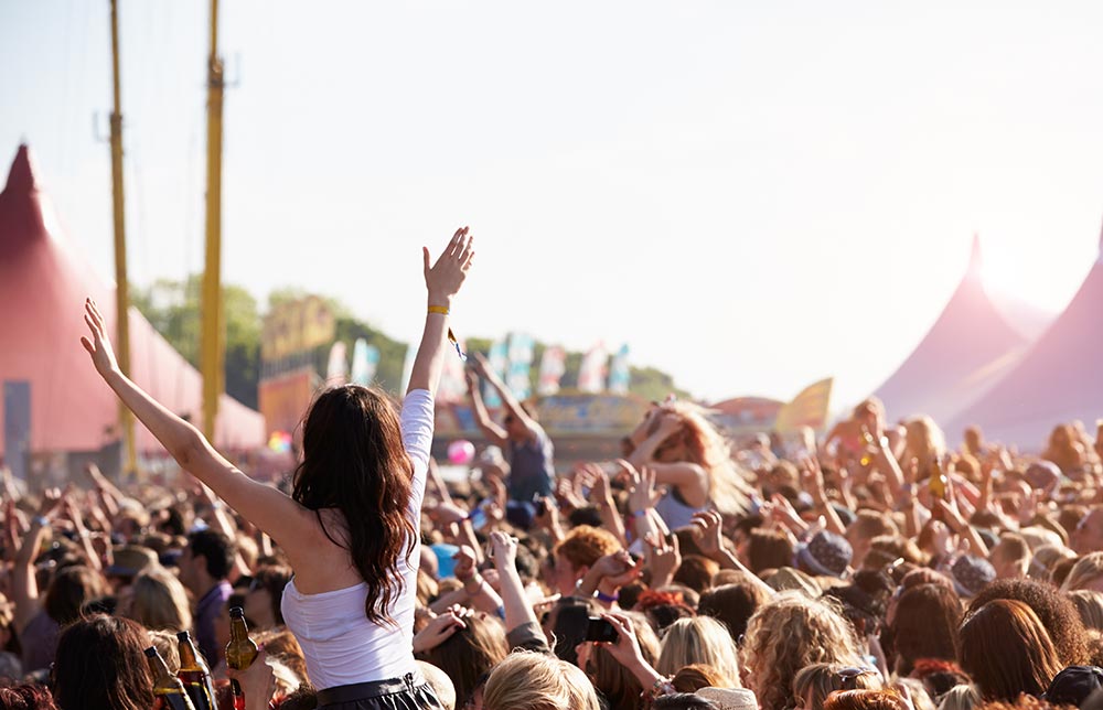 A packed crowd at a music festival watching the stage. A girl sits on top of someone's shoulders with her arms up in the air.