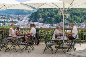 Diners sit at outdoor tables under umbrellas overlooking the city of Salzburg.