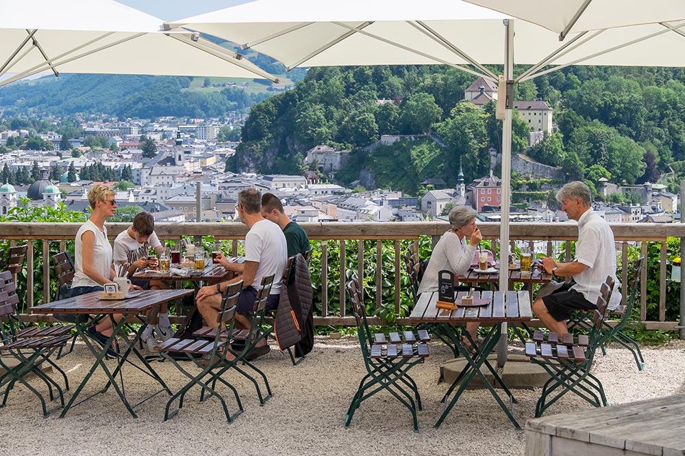 Diners sit at outdoor tables under umbrellas overlooking the city of Salzburg.