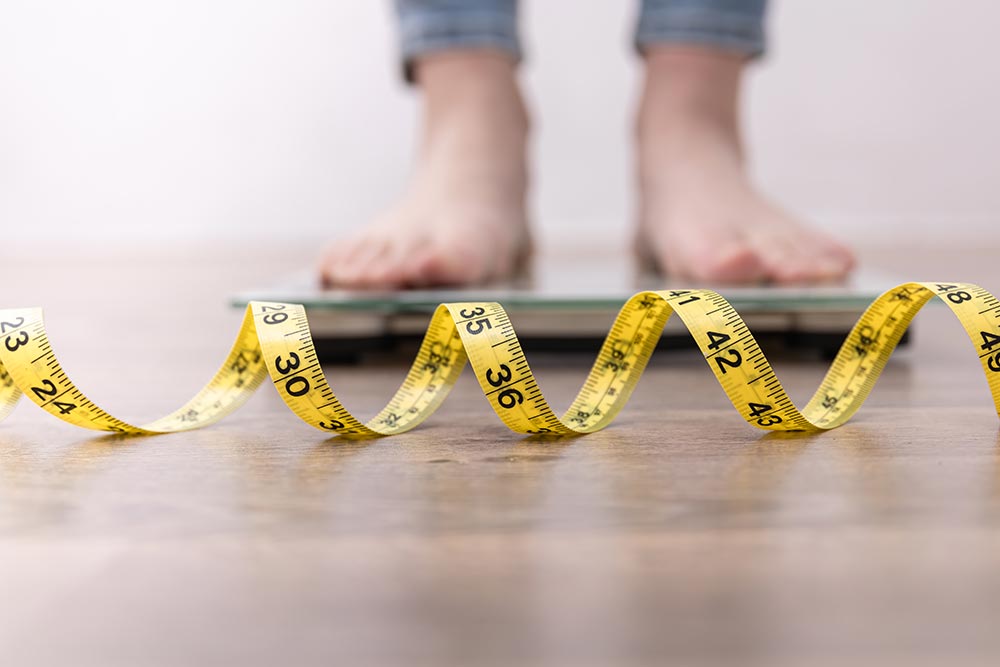 A measuring tape lays on the floor with a person blurred in the background standing on a set of scales.