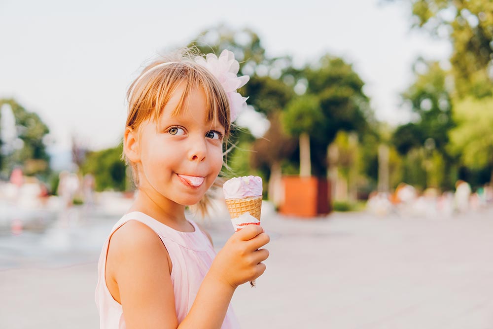 A candid shot of a young girl outside eating an ice cream cone, her tongue slightly poking out and covered with ice cream while she eats. 