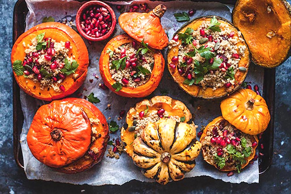 An overhead shot of six different size and colour pumpkins on an oven tray with their lids to the side to reveal a stuffing of quinoa and pomegranate.