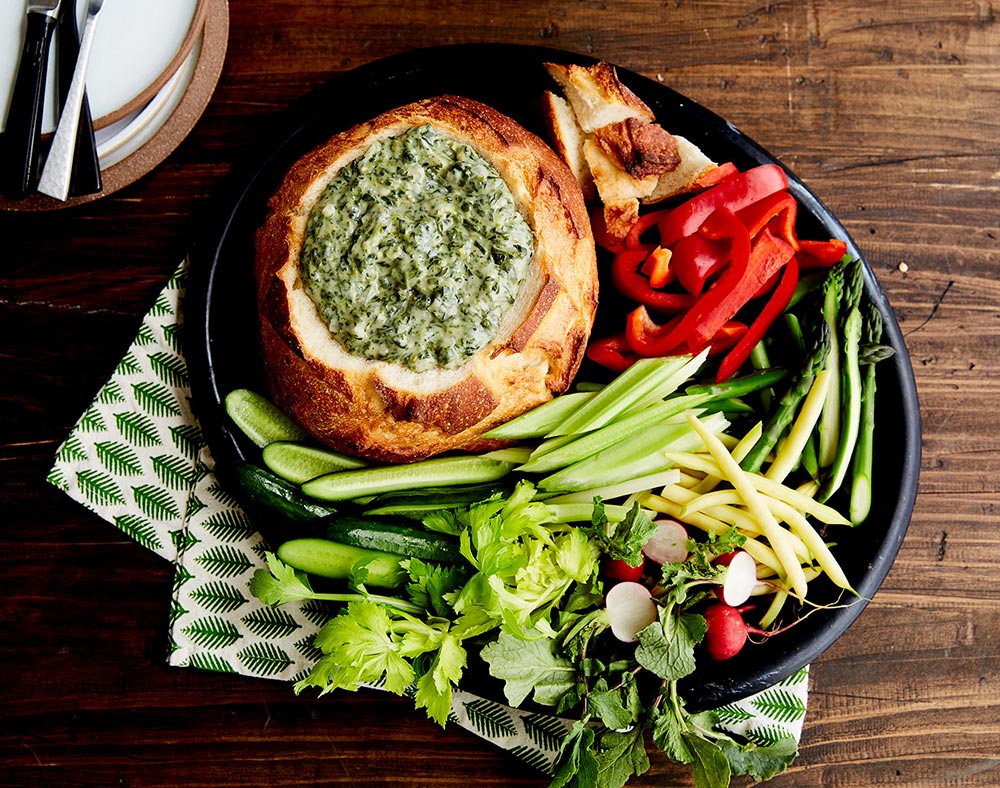 A creamy spinach dip is placed in a cob loaf bowl, surrounded by fresh vegetables for dipping.