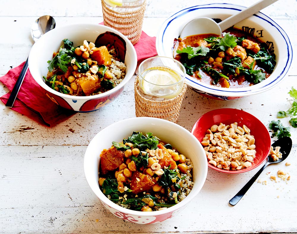 Three bowls of colourful curry sit on a wooden table, garnished with crunchy peanuts.