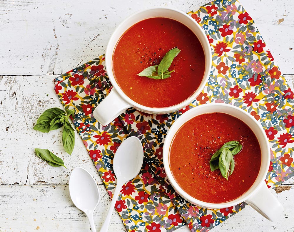 Two bowls of bright red tomato soup sit on a colourful floral placemat on a white bench.