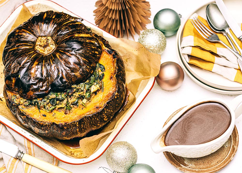 A whole roasted pumpkin sits in an oven dish, with it's lid partially open to reveal a centre filled with stuffing. A gravy boat sits to the side.