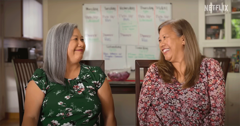 A pair of twins sitting down smiling at each other.