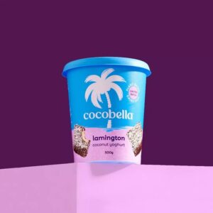 A tub of Cocobella yoghurt in lamington flavour sits in front of a two-tone purple background.