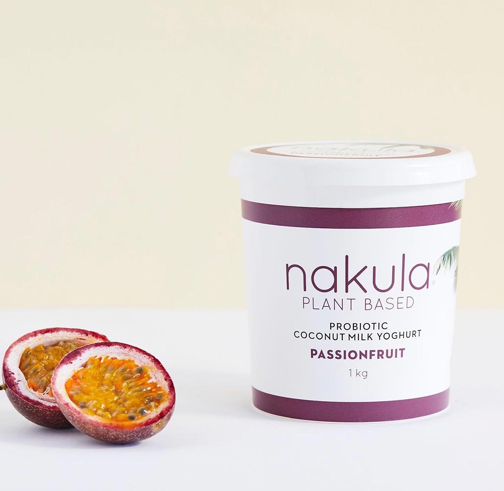 A tub of Nakula passionfruit yoghurt sits on a white surface with two halves of a passionfruit beside it. 