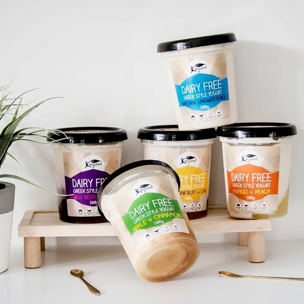 Various flavours of Kingland yoghurt and displayed on a shelf on a bench, with spoons nearby.