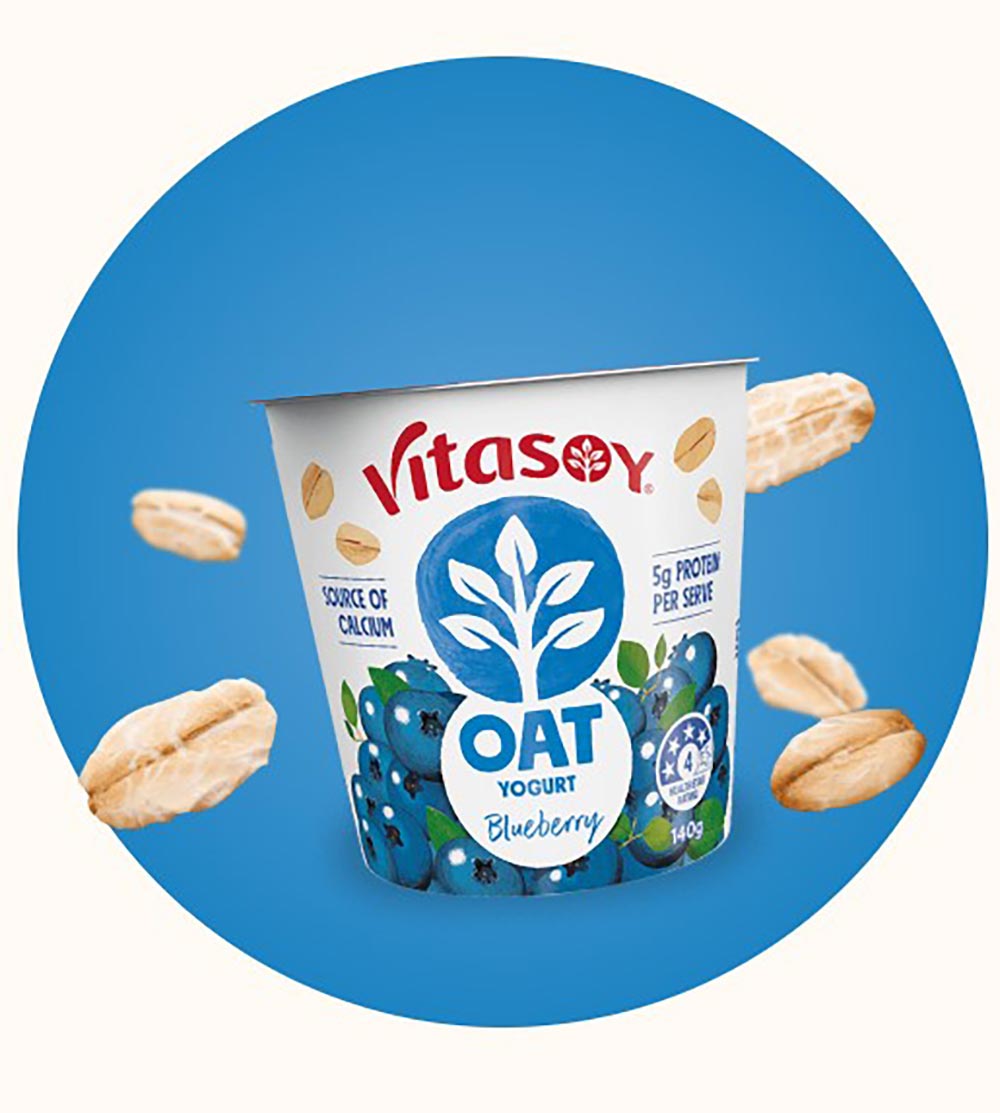 A tub of Vitasoy blueberry oat yoghurt is on a blue circular background. Oats are bursting around the product.