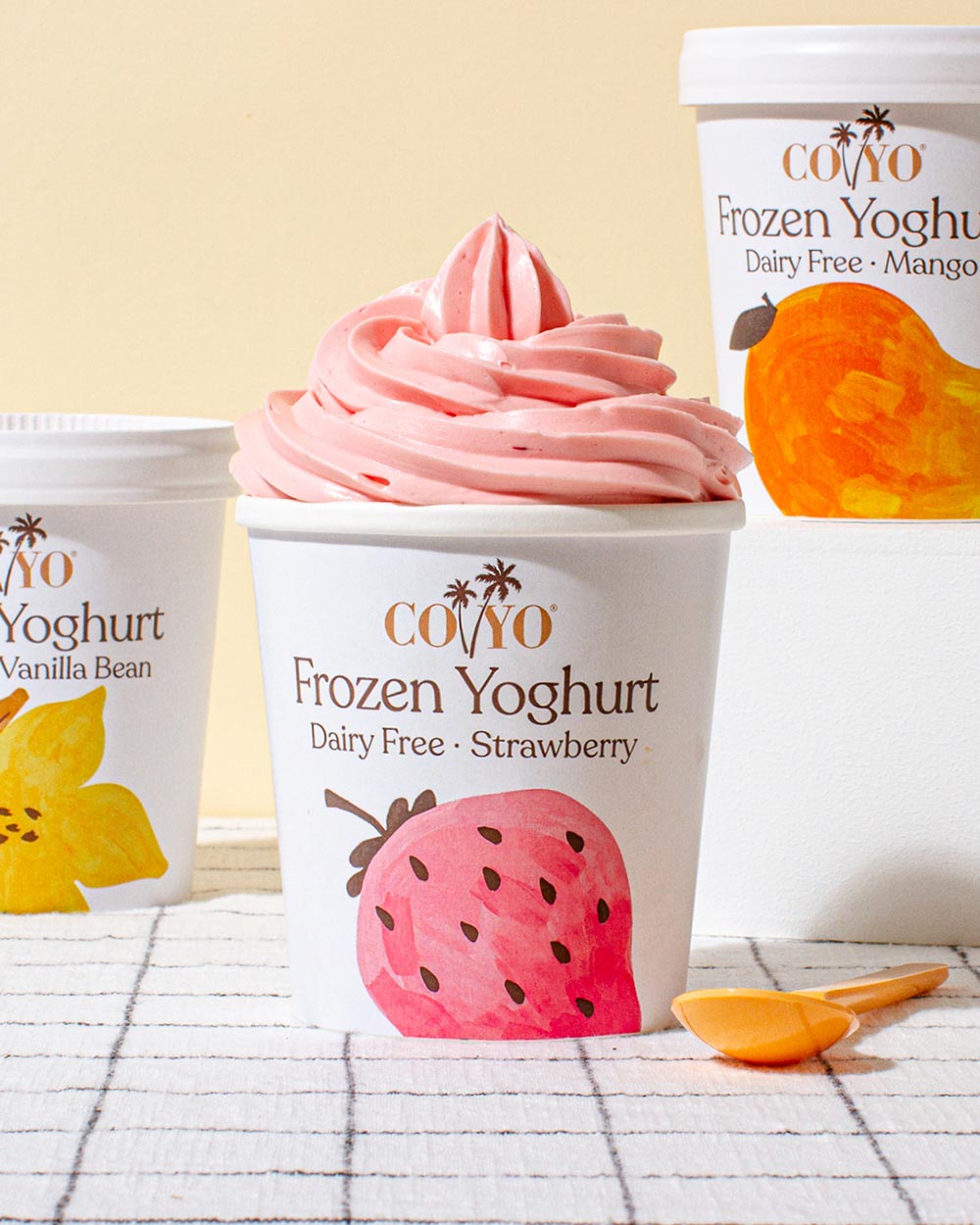 Three tubs of different flavours of COYO frozen yoghurt are on a bench with a peach wall background. The tub in the foreground is open and filled to the brim with strawberry swirls. 