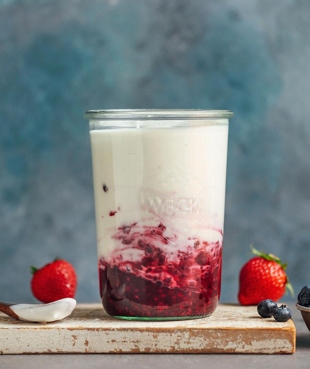 A glass sits on a chopping board filled with berry compote in the lower half and homemade coconut yoghurt.