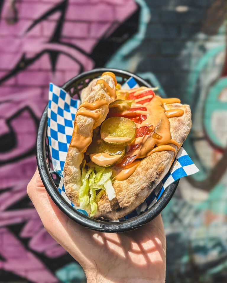 A hand holds up a pizza sandwich hybrid in front of a graffitied wall.