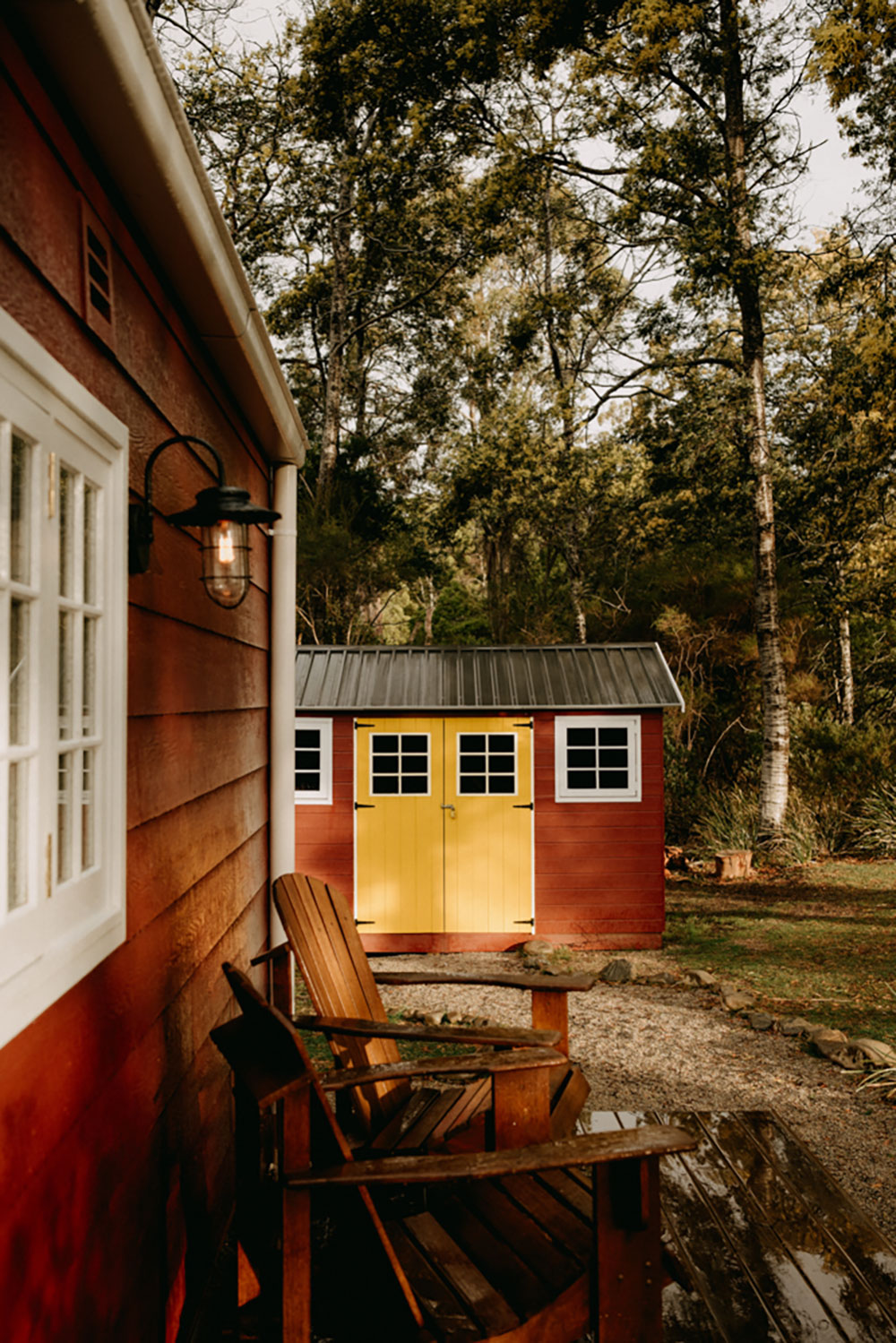 A small red tiny house sits among the trees in rural Tasmania. The doors are a bright yellow, and a fire pit filled with wood sits in the foreground of the shot. 