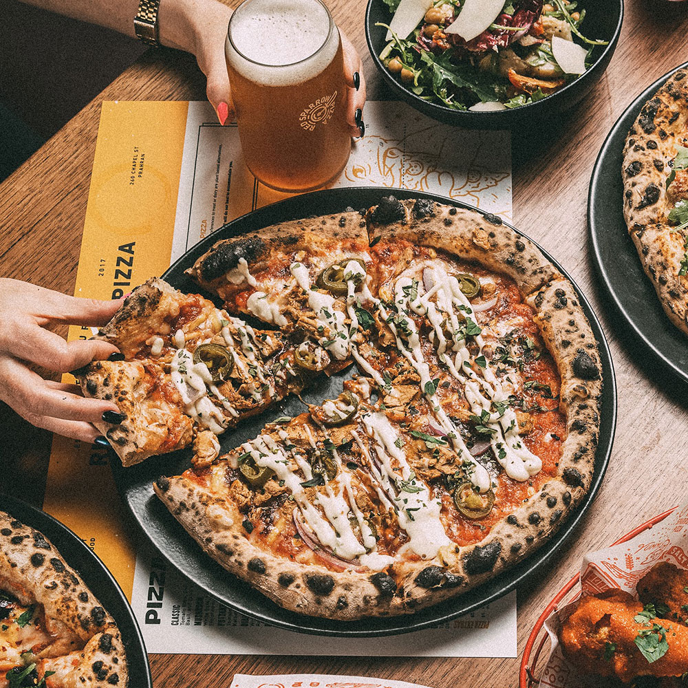 Overhead view of a pizza on a table, with a hand reaching in to grab a slice. The other hand is holding a cold beer. 