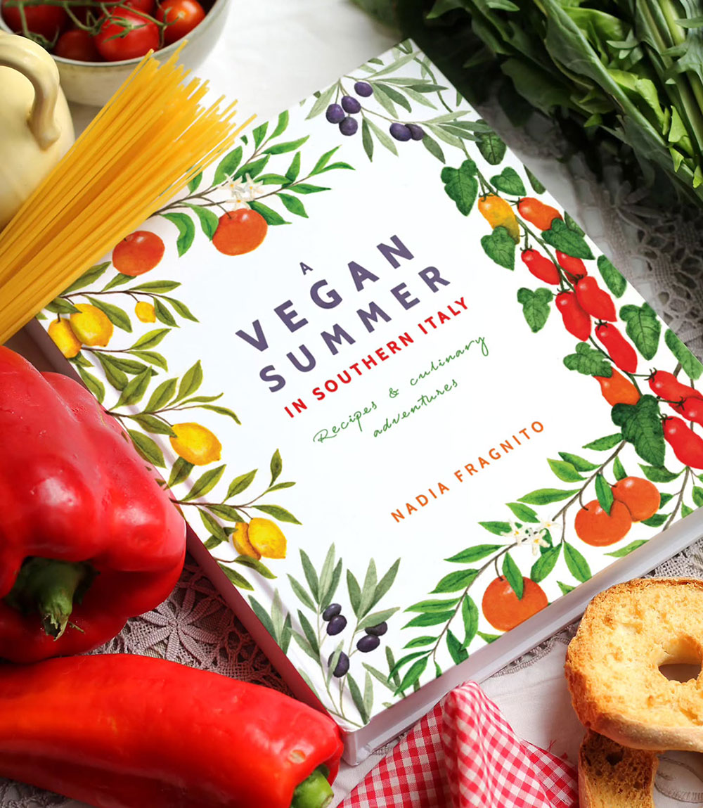 A cookbook, 'A Vegan Summer in Southern Italy' sits on a bench top surrounded by pasta and peppers.