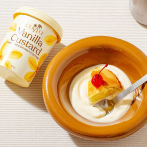 A slice of pie sits floating on custard in a bowl. A tub of COYO Vanilla Custard is shown to the side.