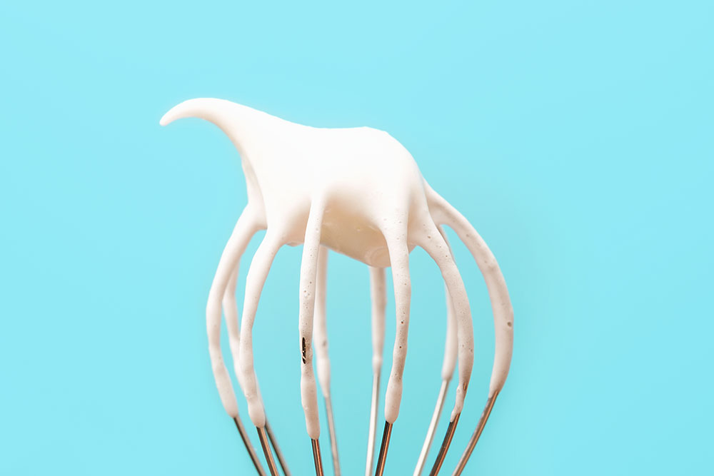 A close-up of a whisk with whipped cream on a blue background.