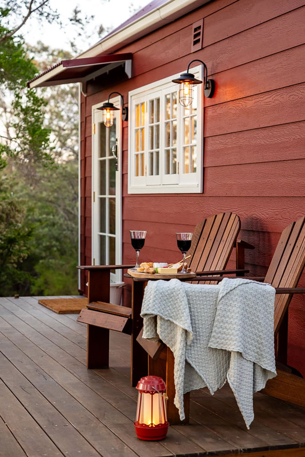 An external view of a tiny red weatherboard house in regional Tasmania. On the front deck sit tit a table setting for two with a vegan charcuterie board and red wine.