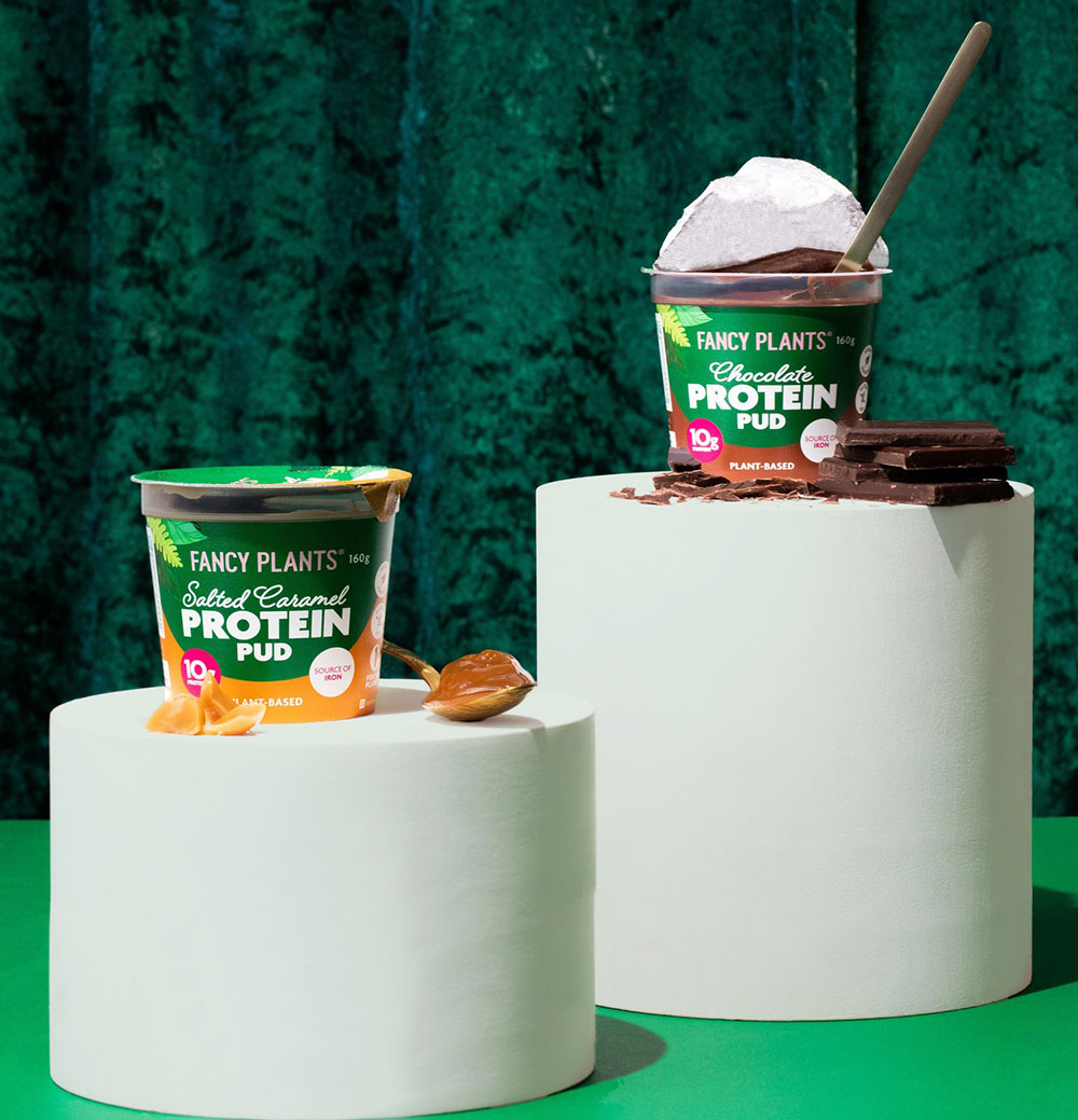 A Salted Caramel and Chocolate Protein Pudding from Fancy Plants are sat atop plinths on a green velvet background.