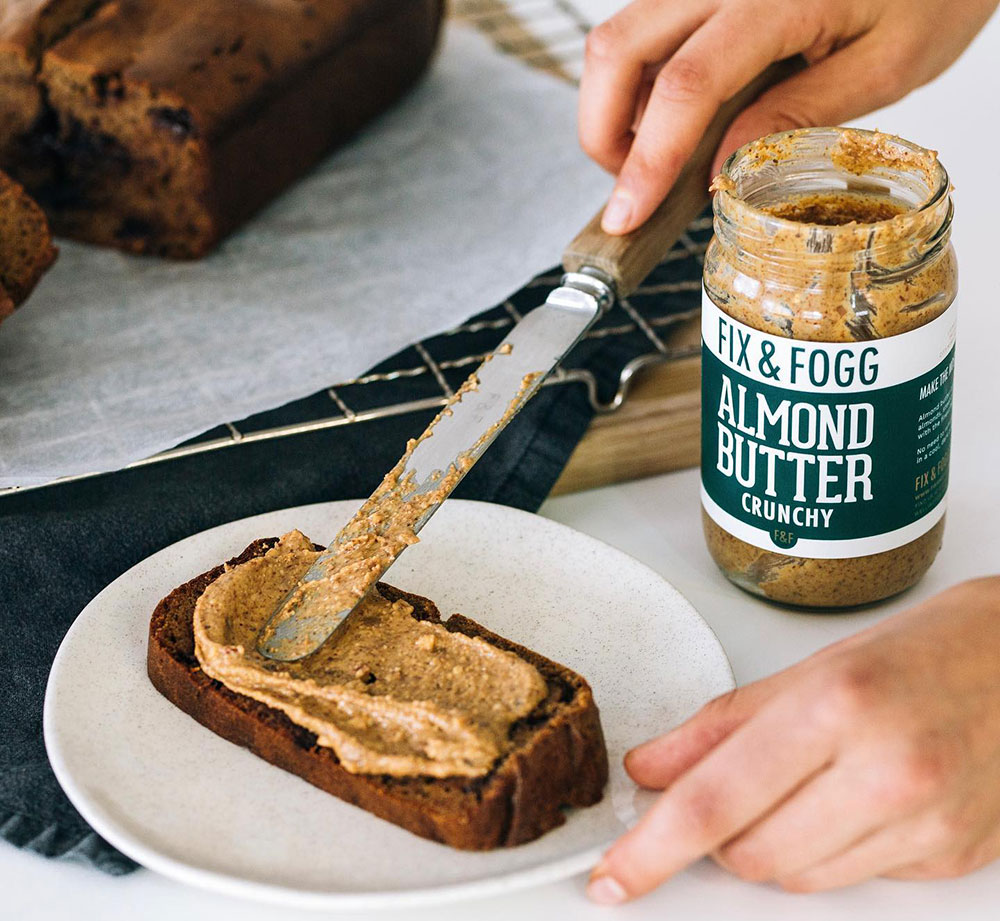 A hand spreads some Fix & Fogg brand almond butter over a slice of banana bread.