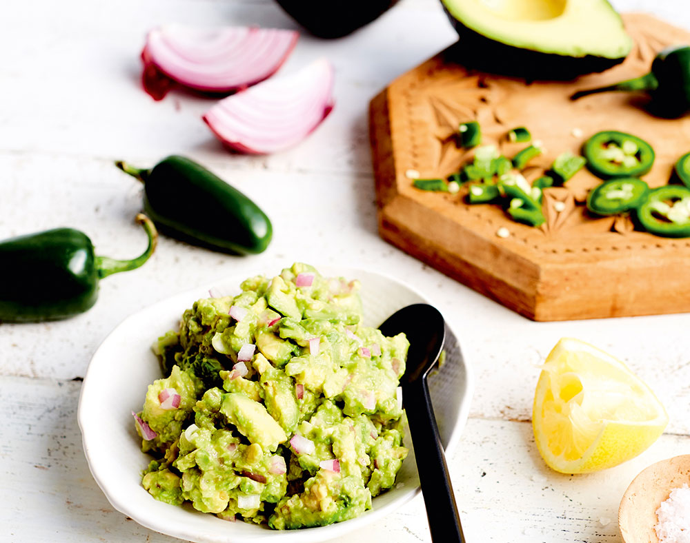 A bowl of guacamole with jalapenos, red onion, and avocado in the background.